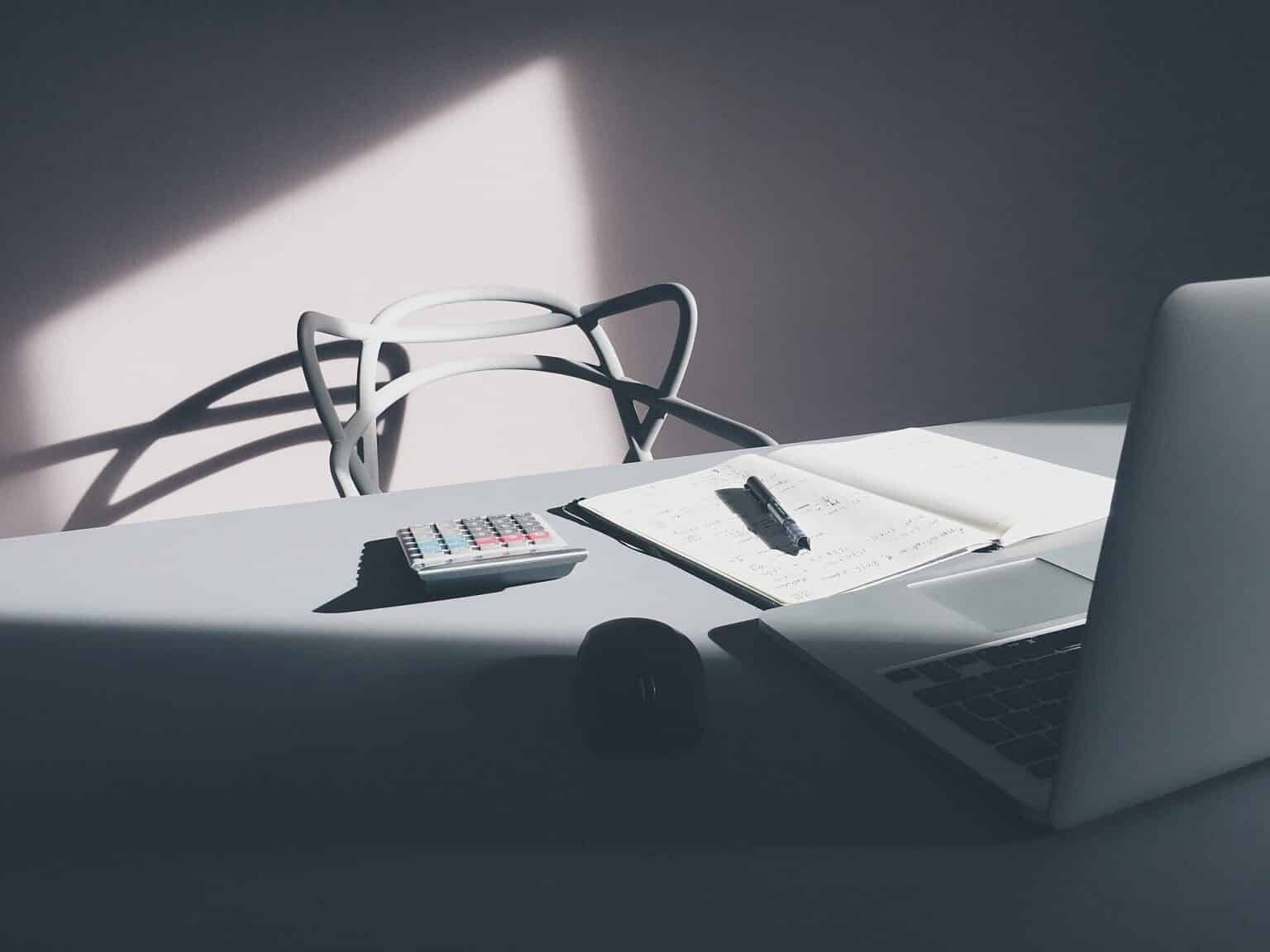 A calculator beside a notebook of handwritten calculations and an open laptop on a sunlit desk