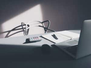 A calculator beside a notebook of handwritten calculations and an open laptop on a sunlit desk